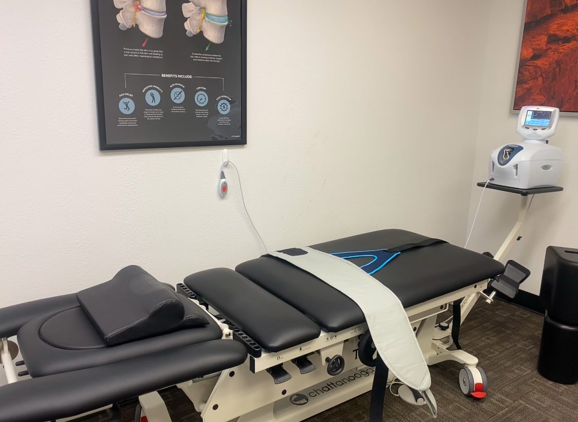 Treatment room with motorized decompression table, wall-mounted educational poster about spinal therapy, and clinical