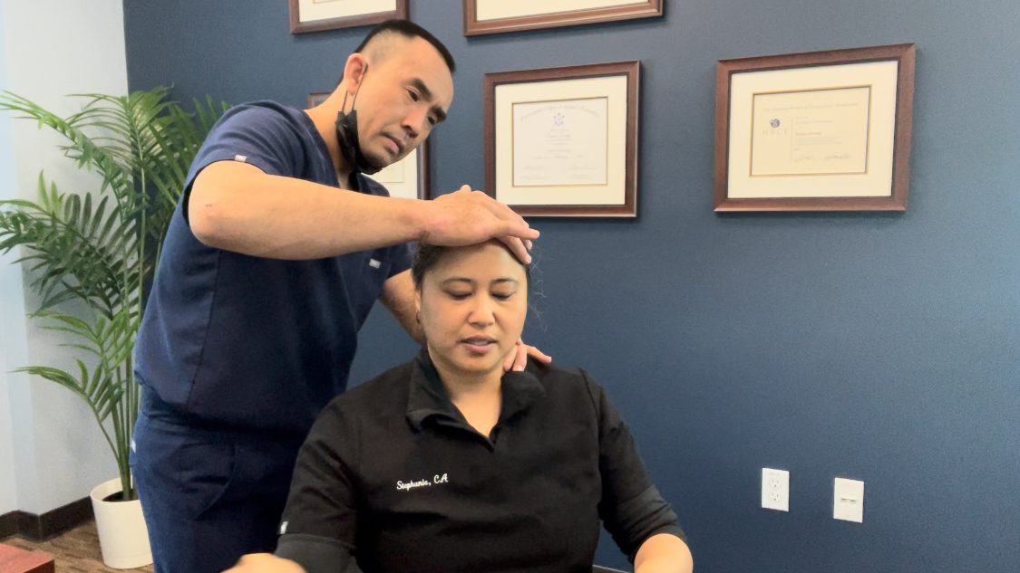 Chiropractor examining patient's head and neck in office with diplomas on wall