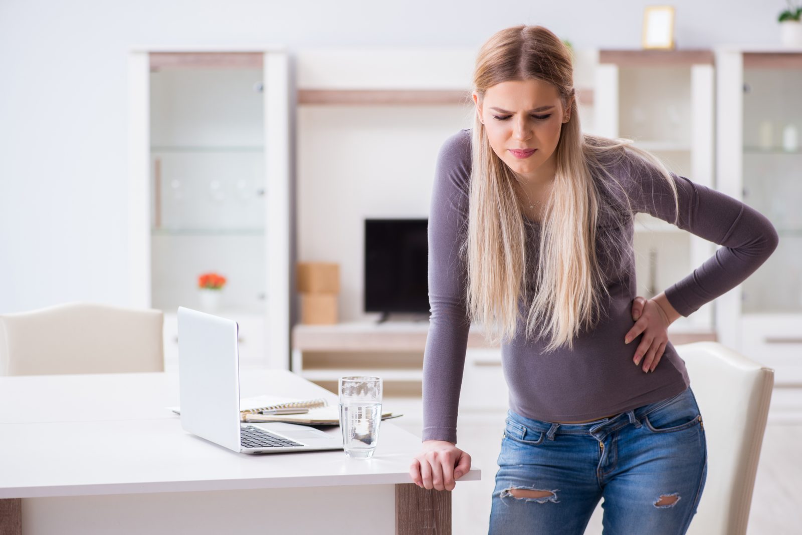 Pregnant woman at desk holding lower back, appearing to experience back pain while working