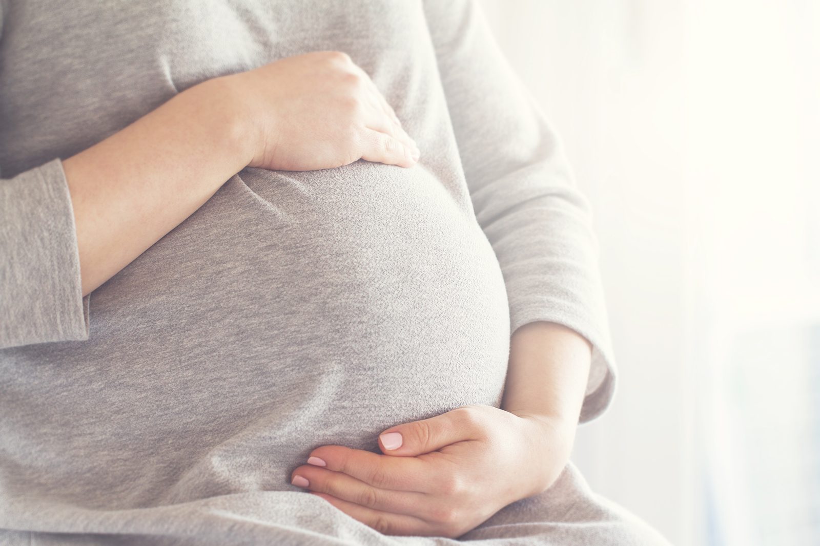 Pregnant woman in gray sweater holding her belly with both hands in a protective gesture
